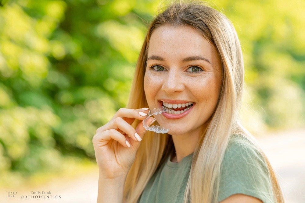 Young woman with long blonde hair smiling while holding a clear aligner outdoors, showcasing Invisalign treatment after understanding about Does Insurance Cover Invisalign in Chester Springs, PA.