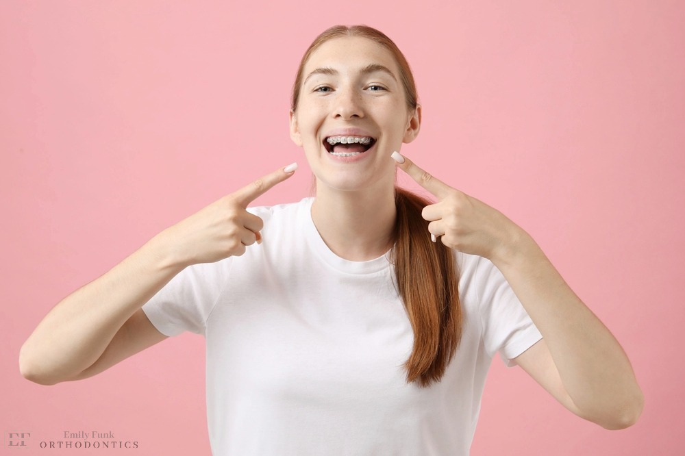 girl pointing her metal braces pink background - Clear Braces vs Metal Braces in Chester Springs, PA